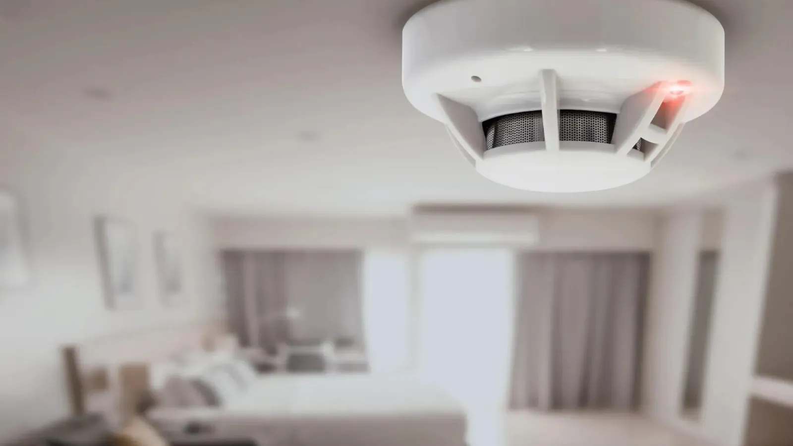 White smoke detector mounted on a bedroom ceiling with blurred bed and window in the background.