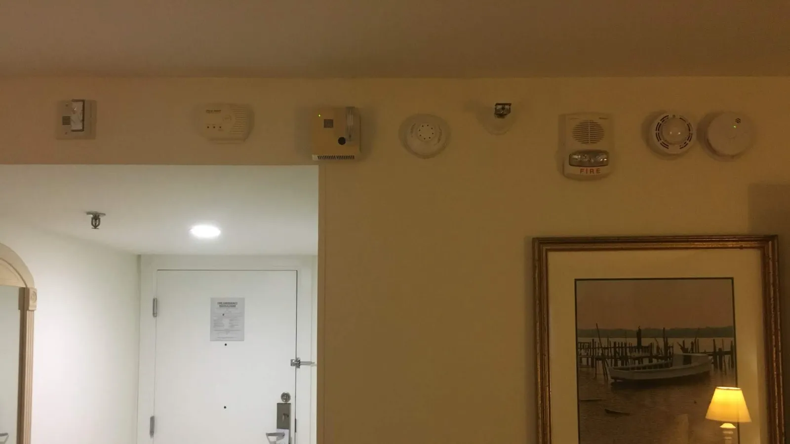 White hotel room door with multiple safety alarms and a framed picture on the wall with a lamp reflection