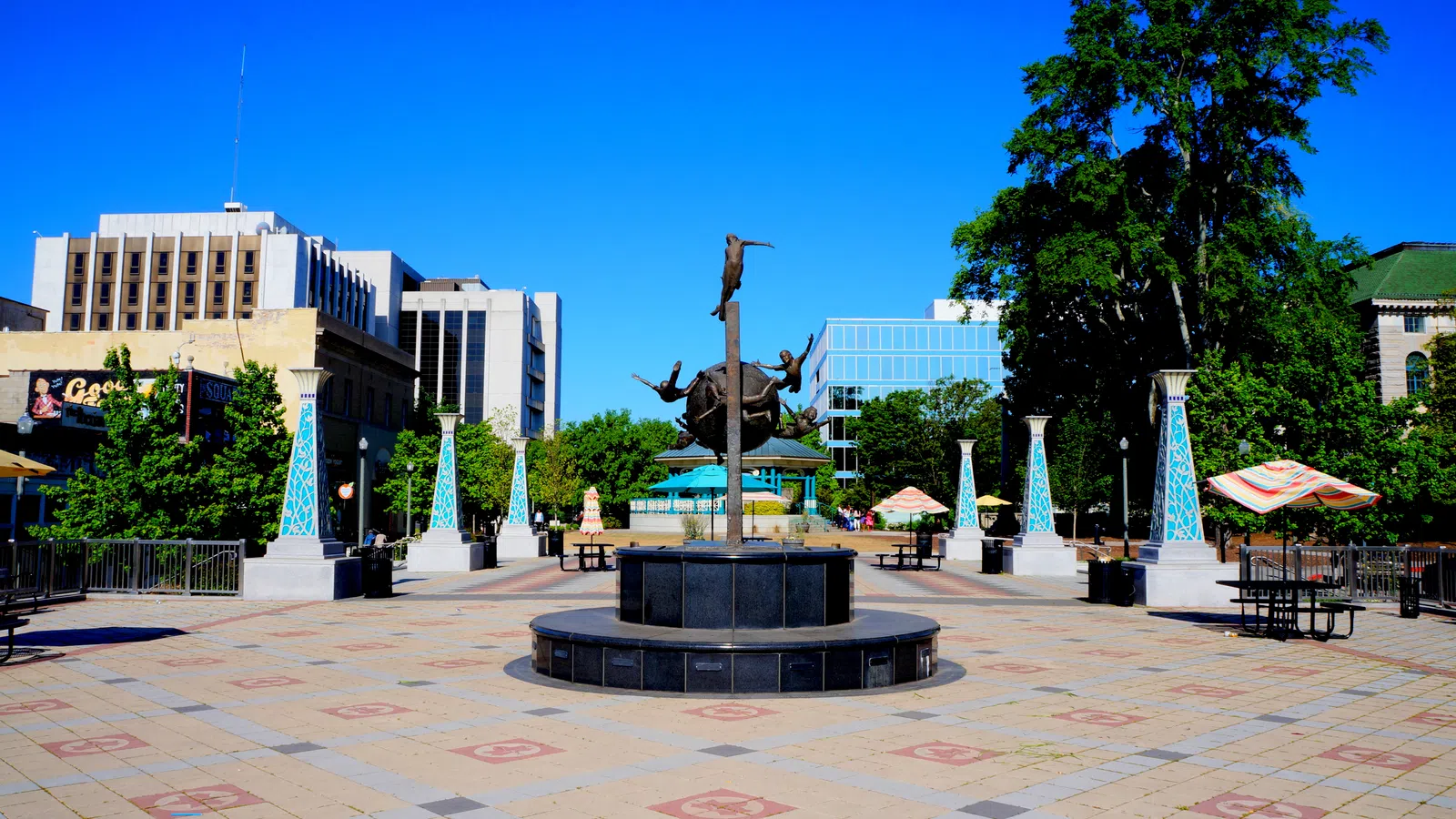 Urban plaza with modern sculpture center, surrounded by benches, umbrellas, trees, and office buildings.