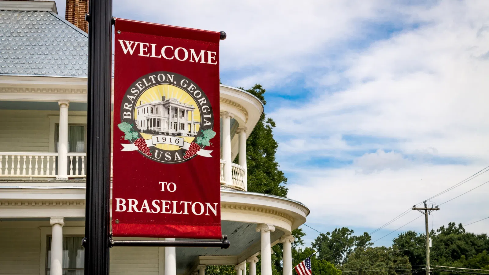 Red welcome sign for Braselton, Georgia hanging from lamppost in front of historic white building with columns.