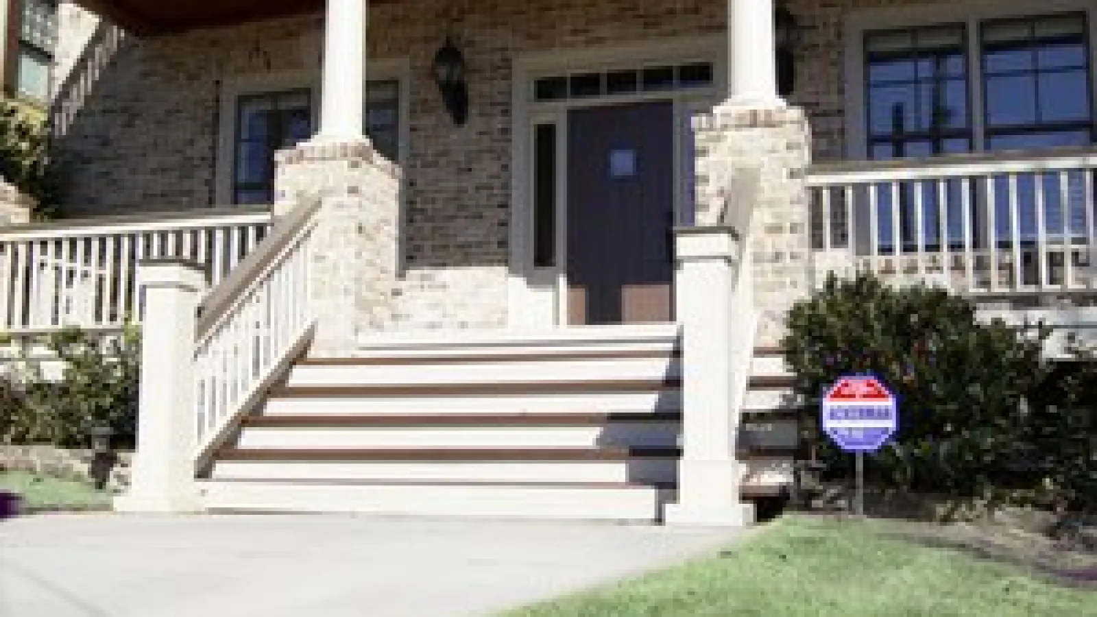 Brick house entrance with white columns, wooden stairs, railing, front door, and security system sign on lawn