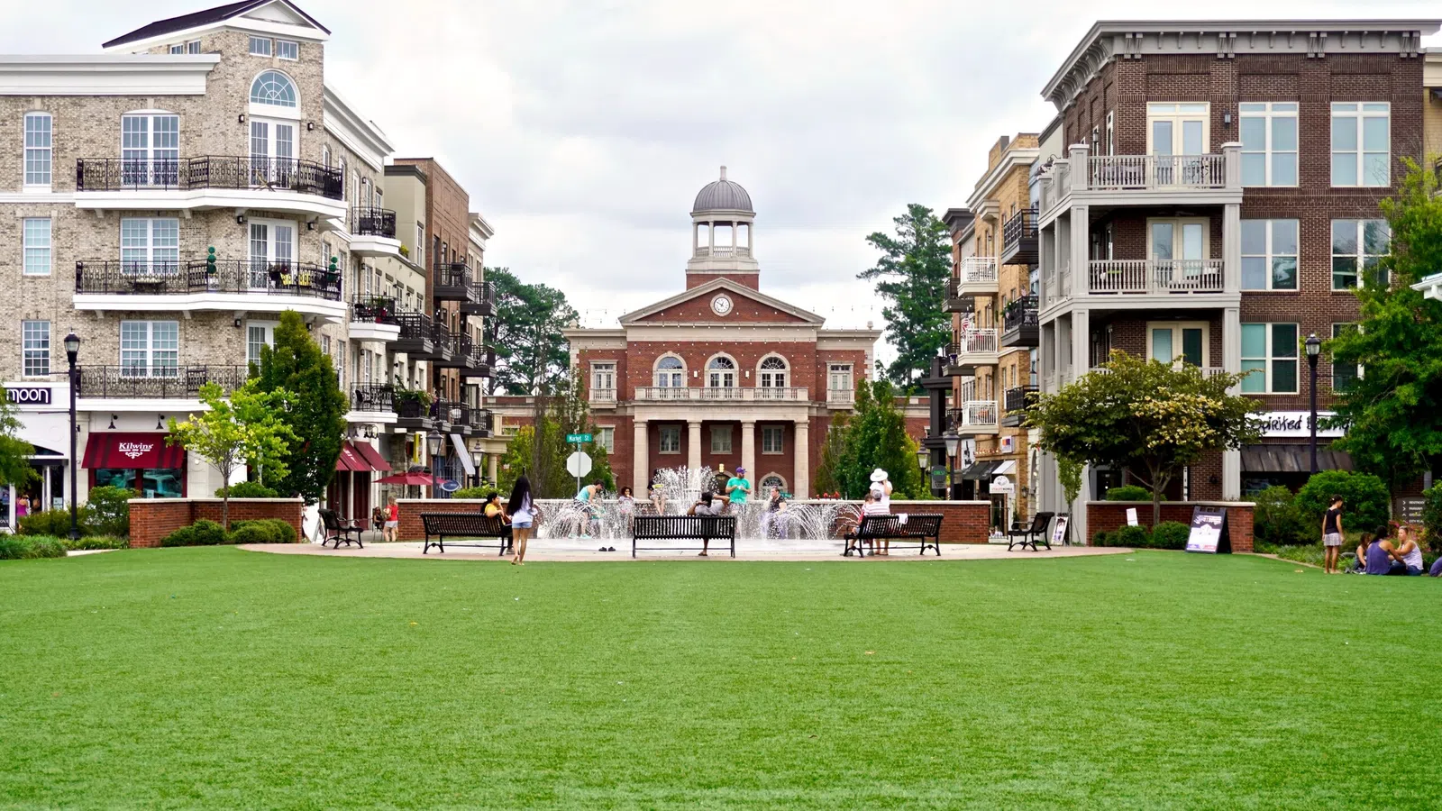 Green park with fountain and benches surrounded by multi-story brick buildings and a central historic clock tower.