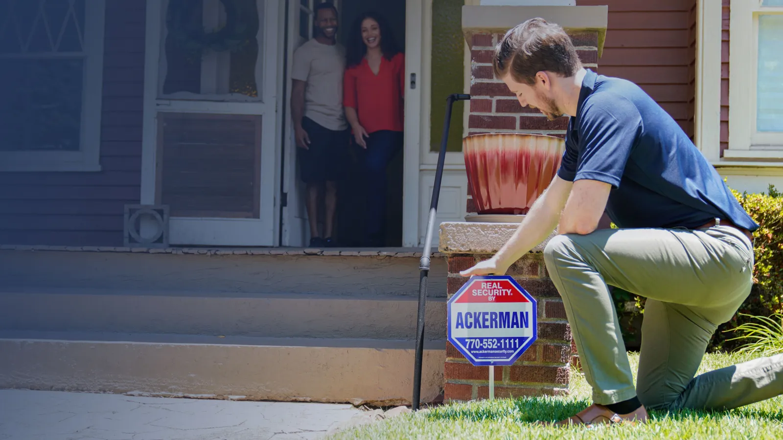 Man installing Ackerman real security sign in front yard as couple watches from doorstep