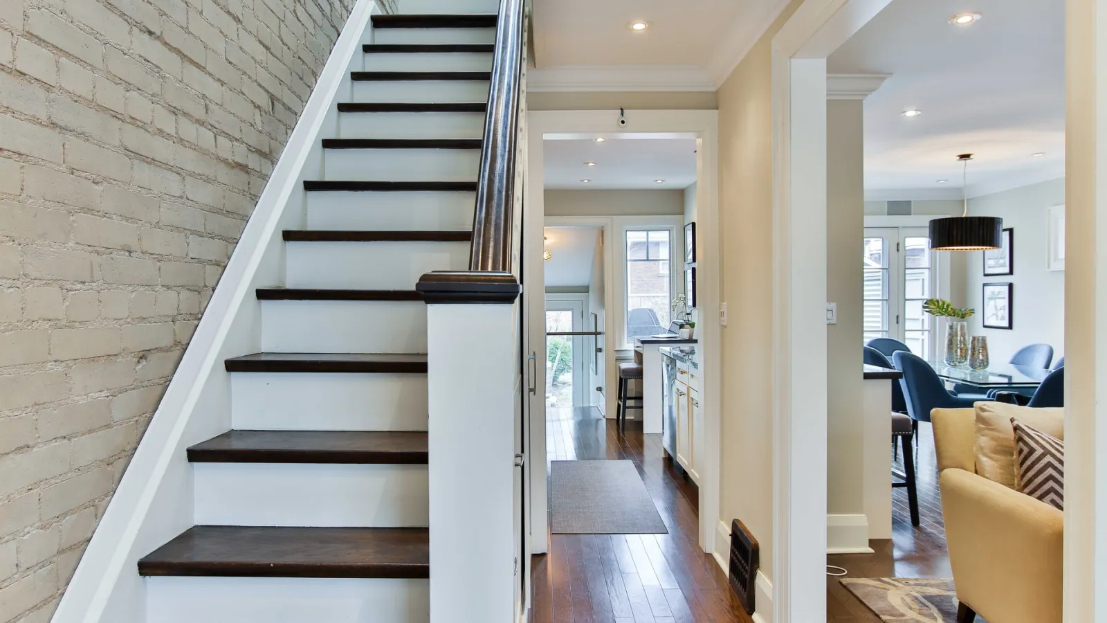 Modern interior with wooden stairs, hardwood floors, kitchen hallway, and living room with beige sofa and dining area.