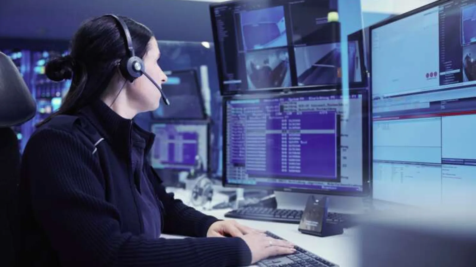 Woman wearing headset working on multiple monitors with data and security footage in a control room.