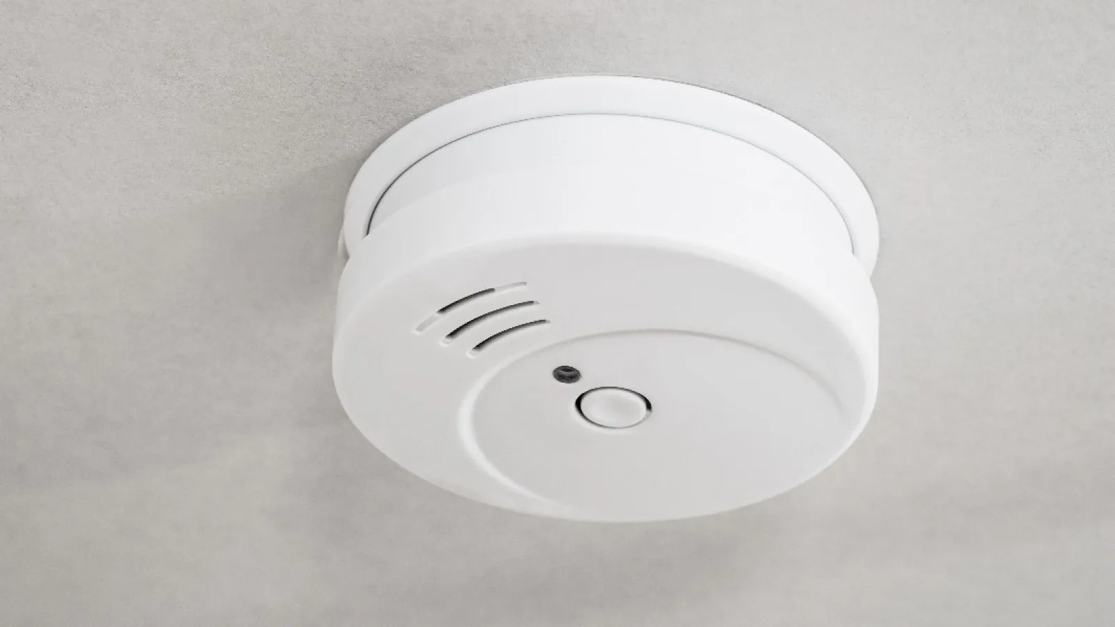 White smoke detector mounted on ceiling with test button and sensor hole visible in close-up