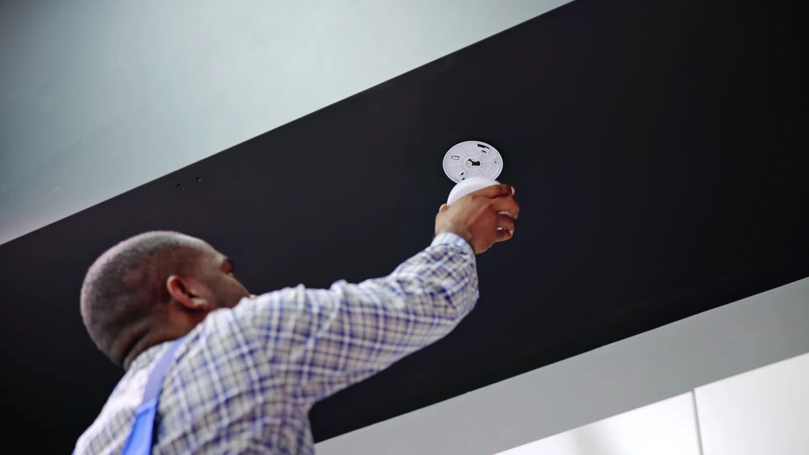 Technician installing a smoke detector on a black ceiling in a modern building interior.