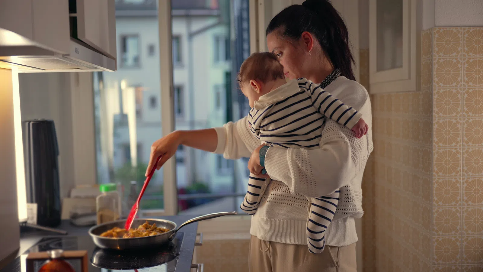 Mother holding baby while stirring food in a pan in a modern kitchen near a large window with daylight.