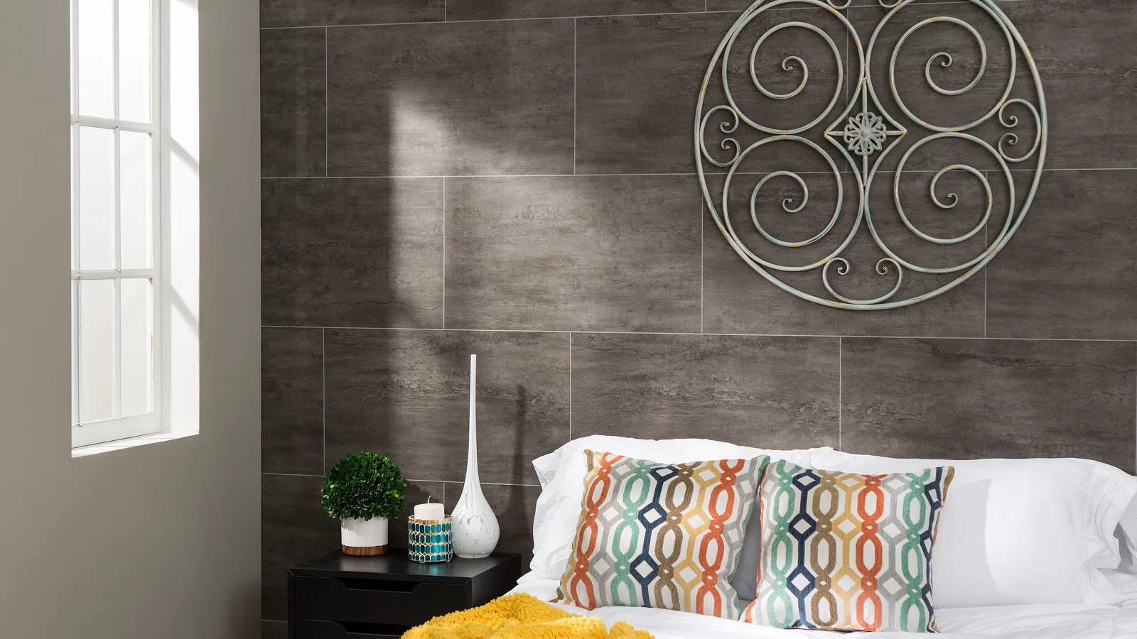 Modern bedroom with gray tile wall, colorful patterned pillows, yellow throw blanket, and decorative metal wall art.