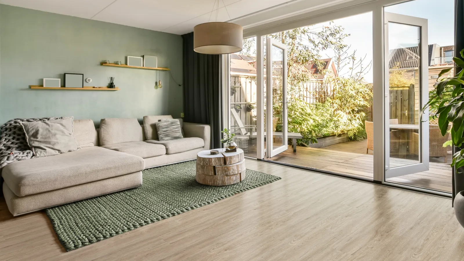 Modern living room with beige sectional sofa, green rug, wooden coffee table, and open glass doors to sunny patio.