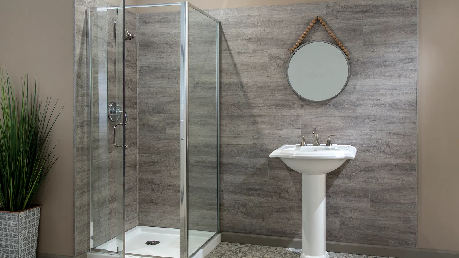 Modern bathroom with glass shower enclosure, pedestal sink, round mirror, and potted plant on tiled floor.