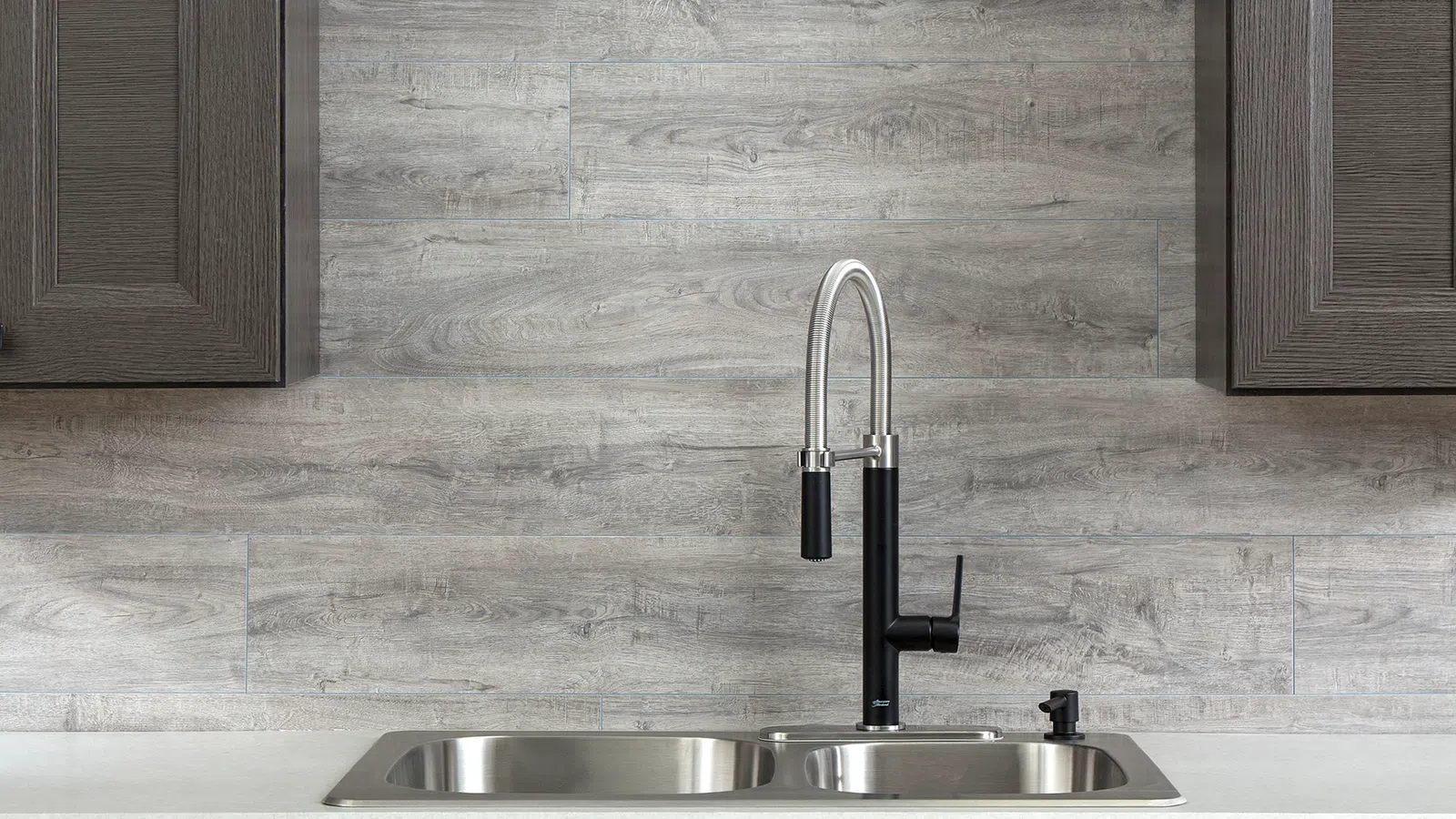 Modern kitchen sink with dual stainless steel basins, black and chrome faucet, gray wood cabinets, and light gray backsplash.