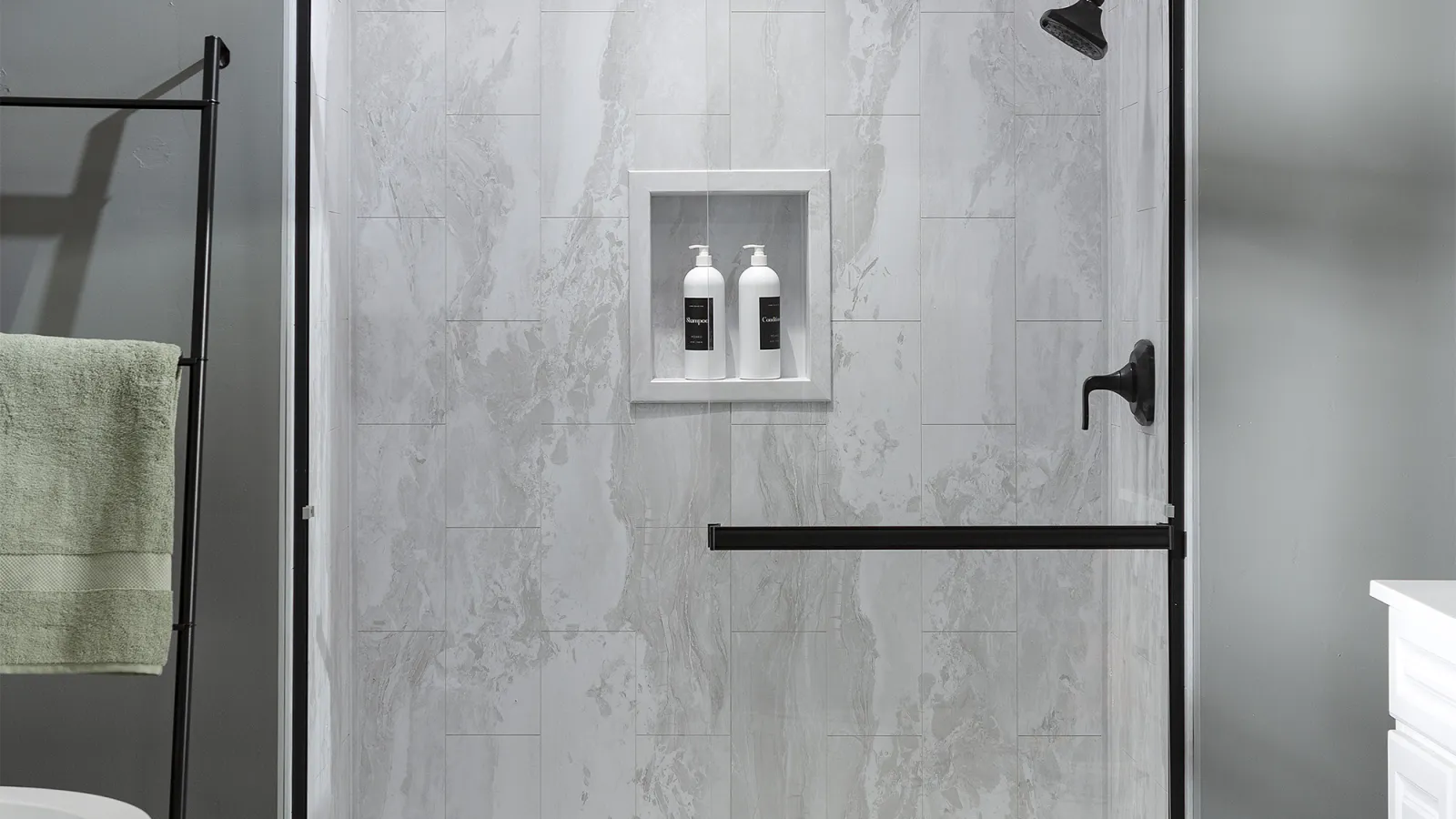 Modern bathroom shower with glass door, gray marble tile walls, black fixtures, and recessed shelf with bottles.