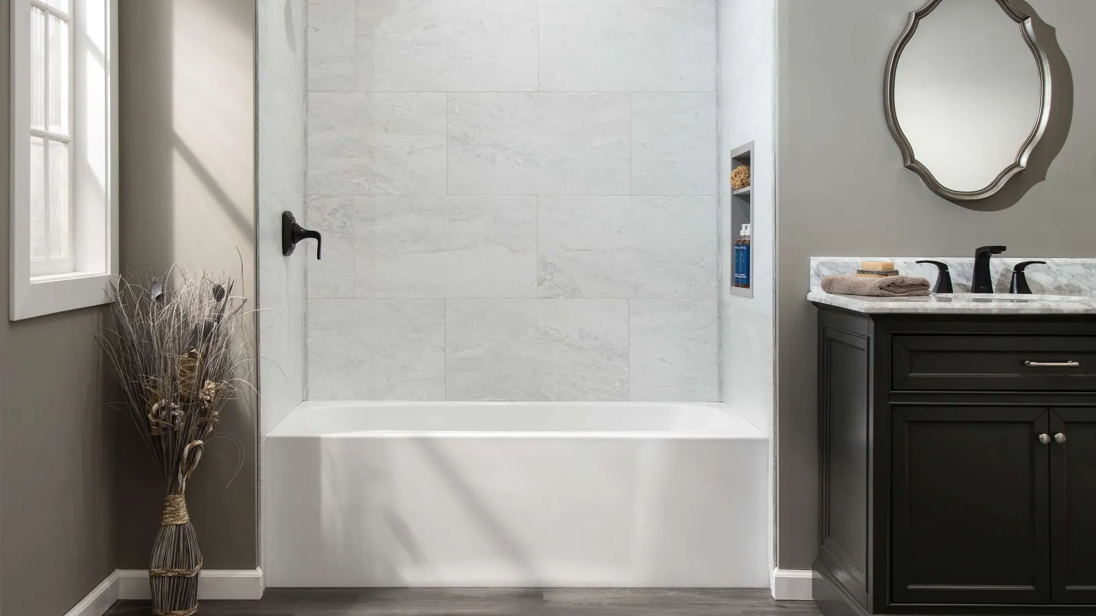 Modern bathroom with white bathtub, gray tiled walls, dark wood vanity, and decorative round mirror under natural light.