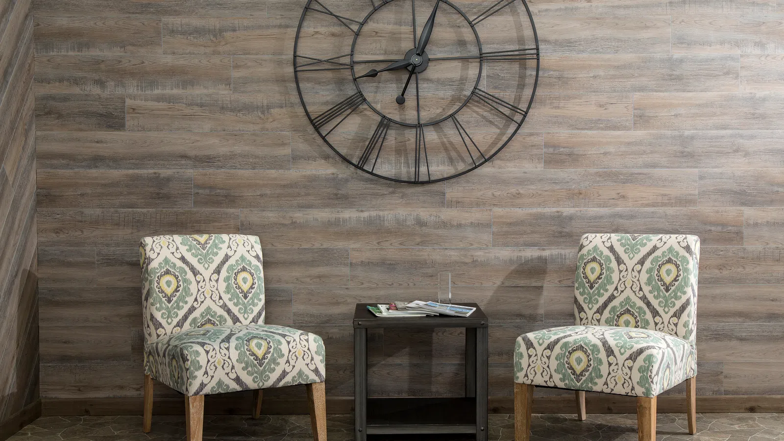 Two patterned armchairs with wooden legs and a small black table below a large black metal wall clock on a wood-paneled wall.
