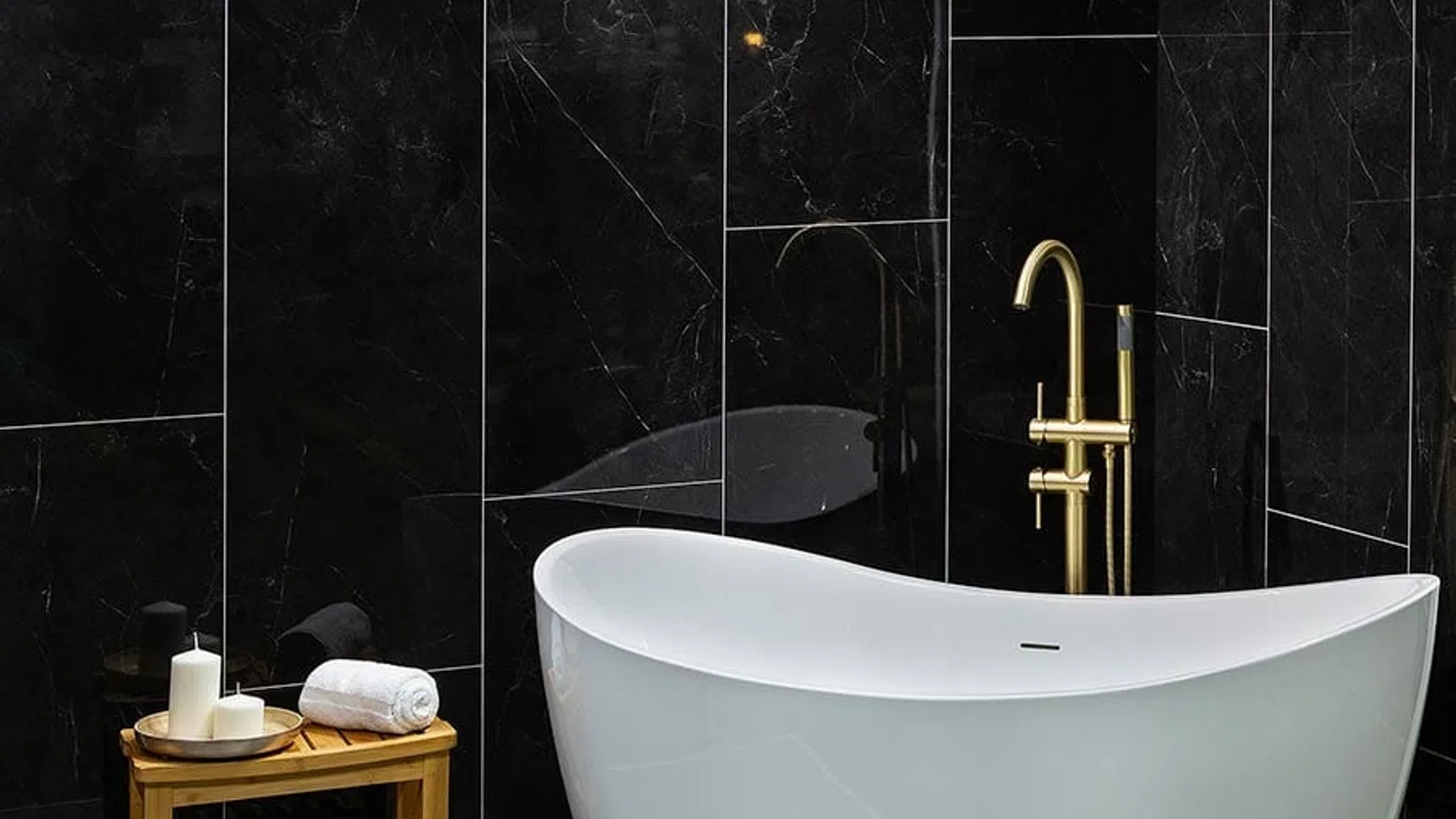 Modern bathroom with white freestanding tub, gold faucet, black marble walls, and wooden side table with candles.