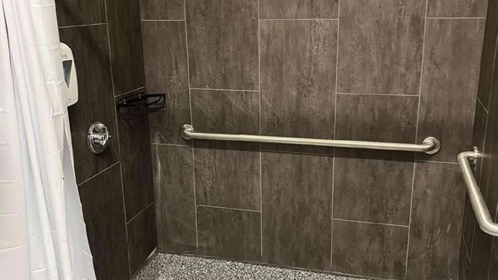 Accessible walk-in shower with gray tiled walls, grab bars, white shower curtain, and textured floor.
