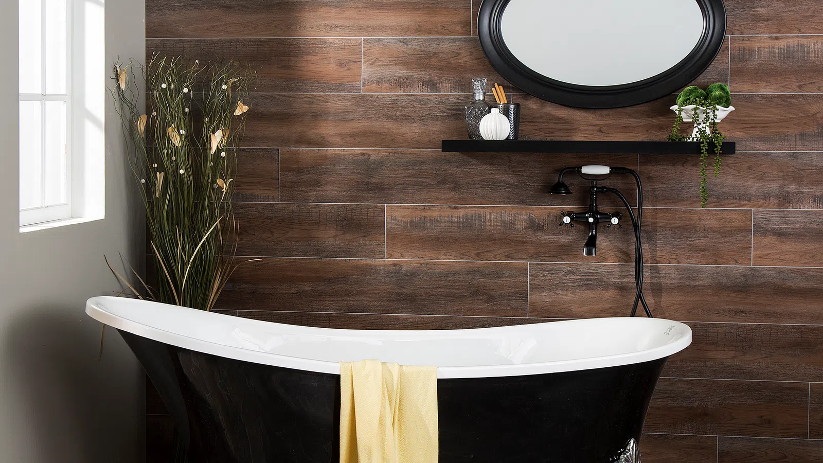 Black clawfoot bathtub with yellow towel, wooden wall tiles, oval mirror, and decorative plants in a bright bathroom.