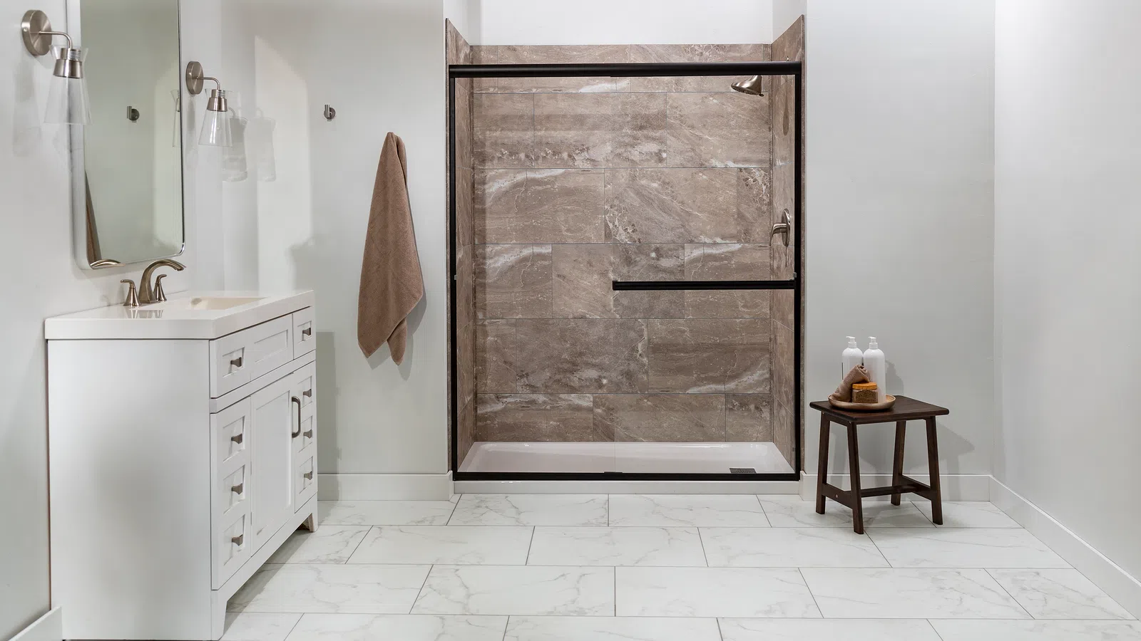 Modern bathroom with white vanity, marble floor, glass shower enclosure, towel, and small wooden stool with toiletries