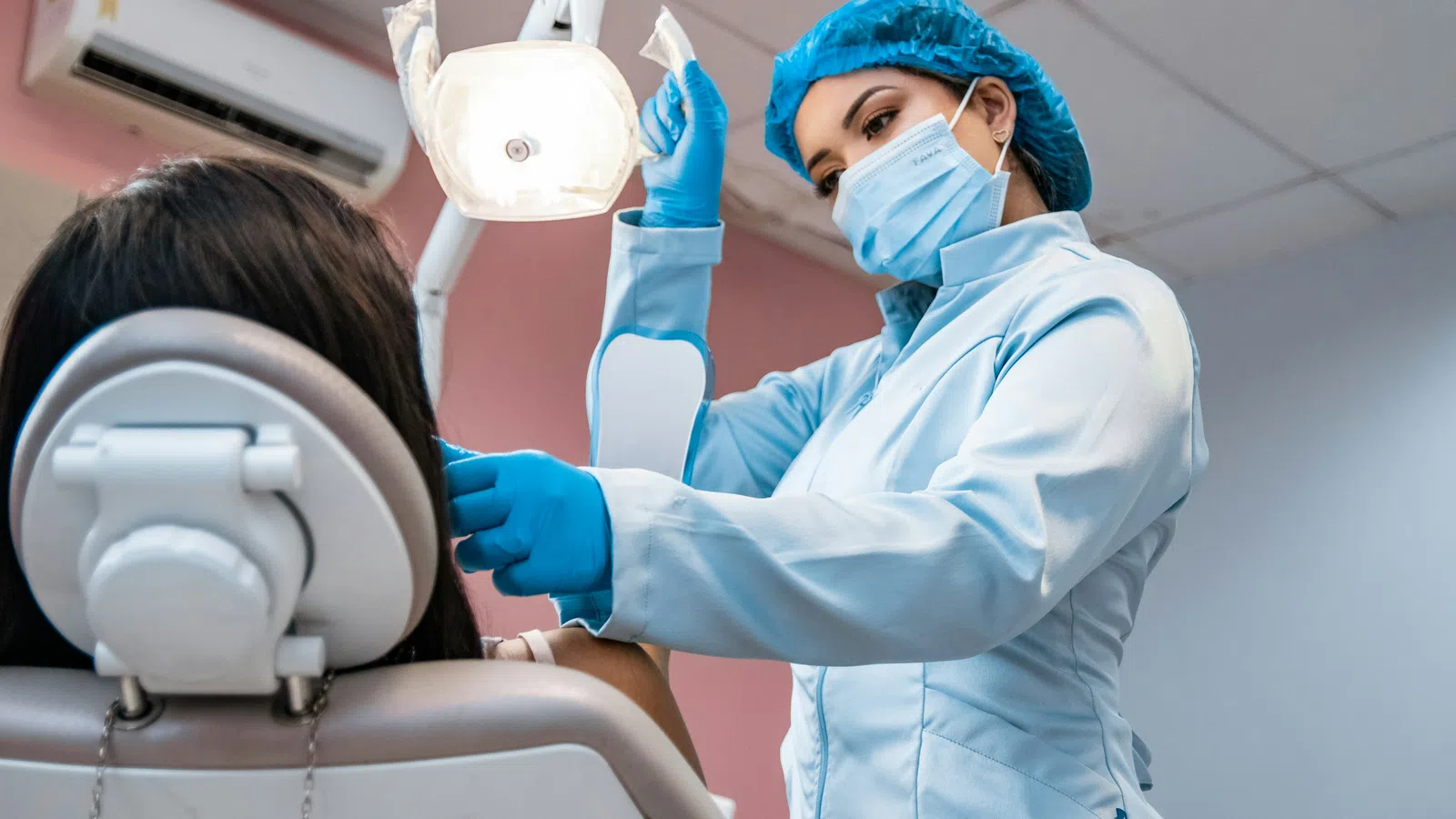Female dentist in protective gear adjusting overhead dental light for patient in clinic chair