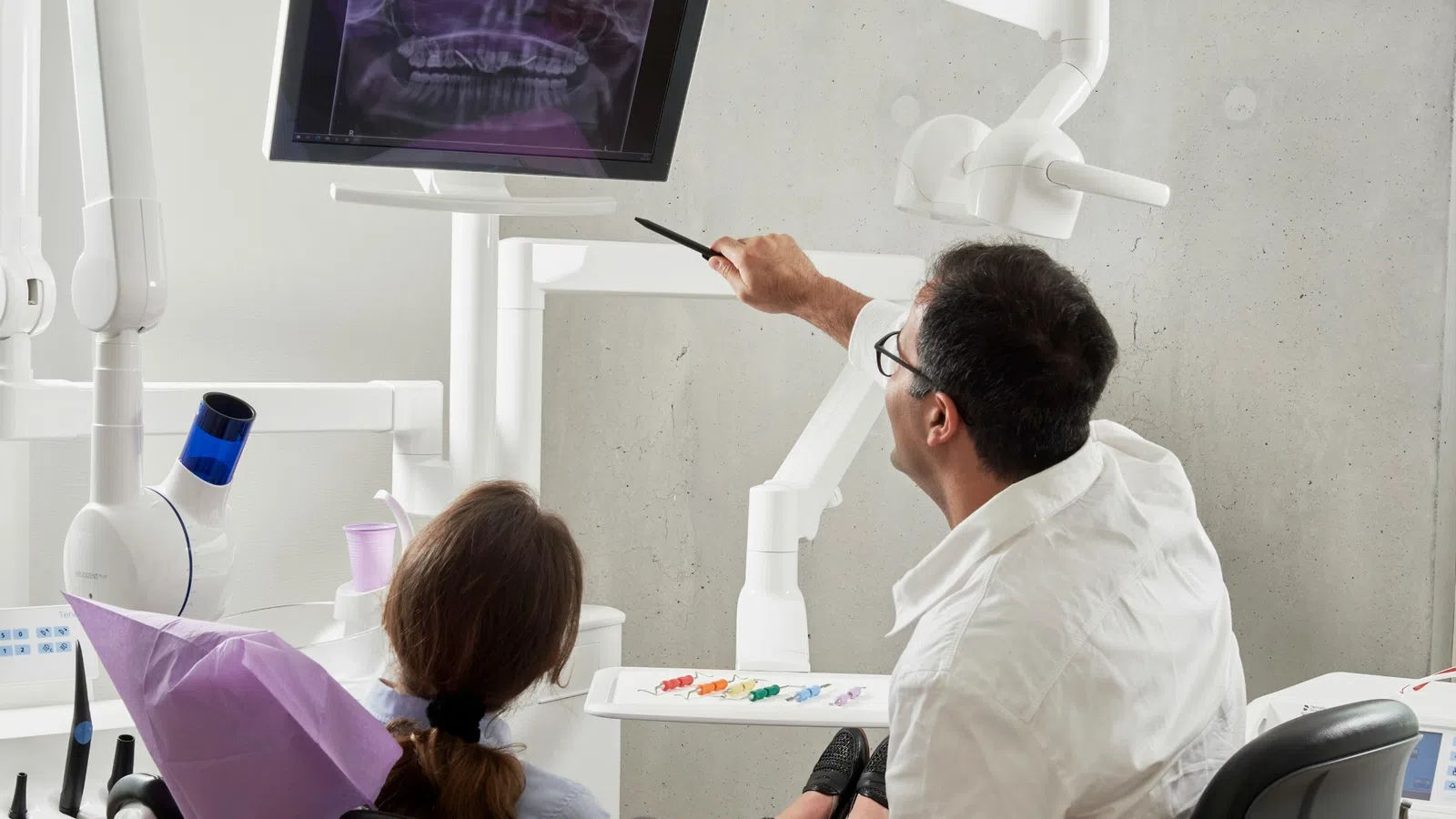 Dentist explaining dental X-ray results to patient seated in clinic chair under bright light.