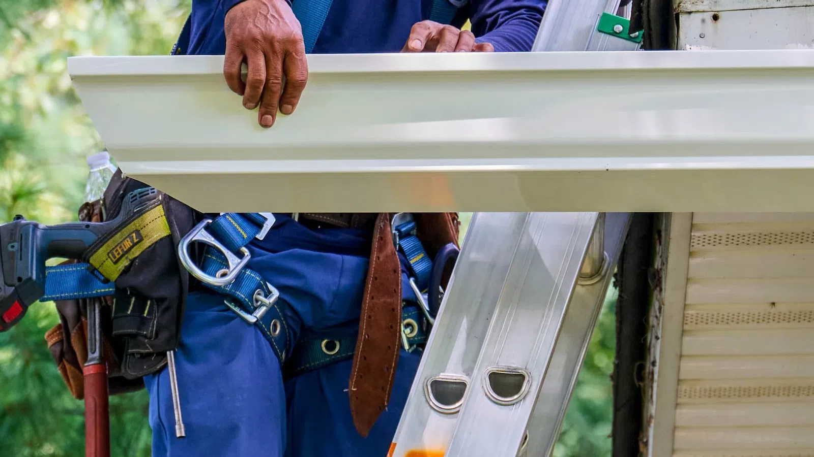 Worker in safety harness holding white gutter on ladder near house eaves for installation
