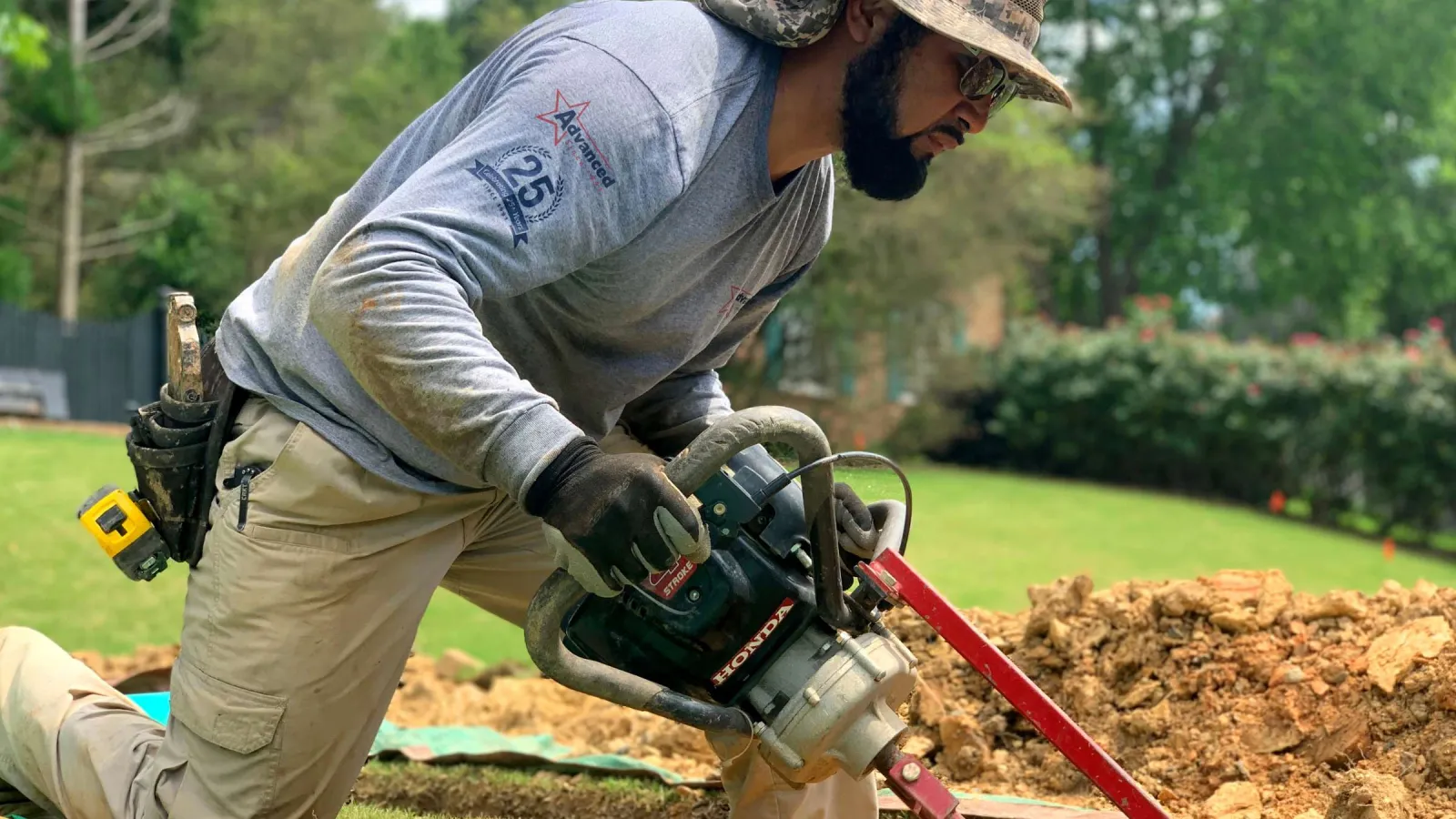 Man wearing a hat and gloves kneeling on grass using a Honda power tool for landscaping work outdoors.