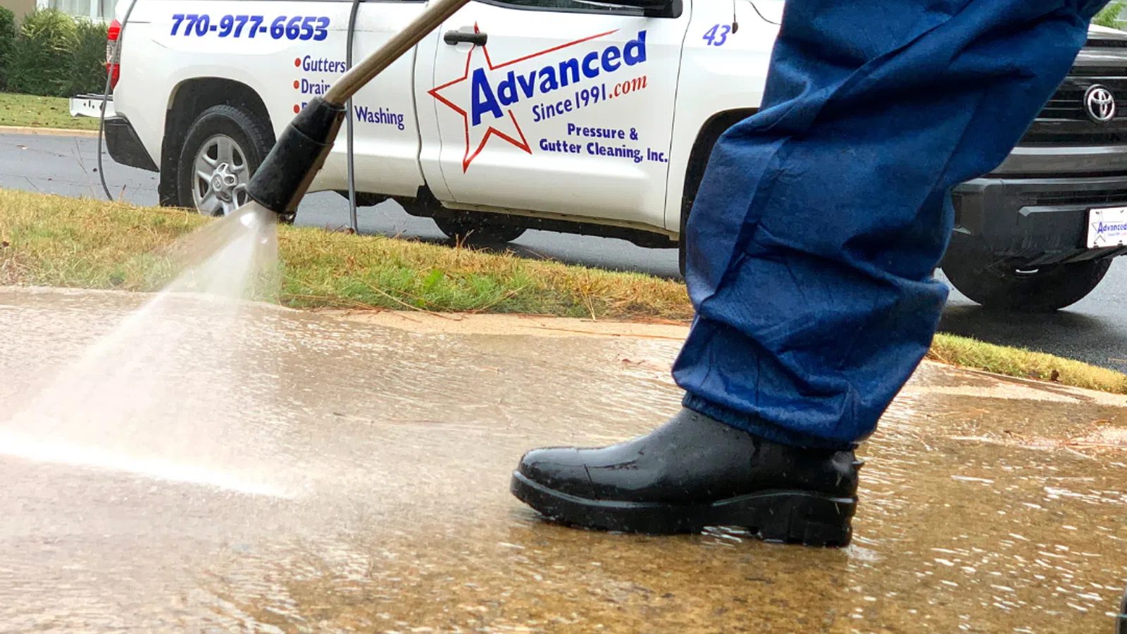 A person in blue overalls pressure washing a driveway next to a service truck.