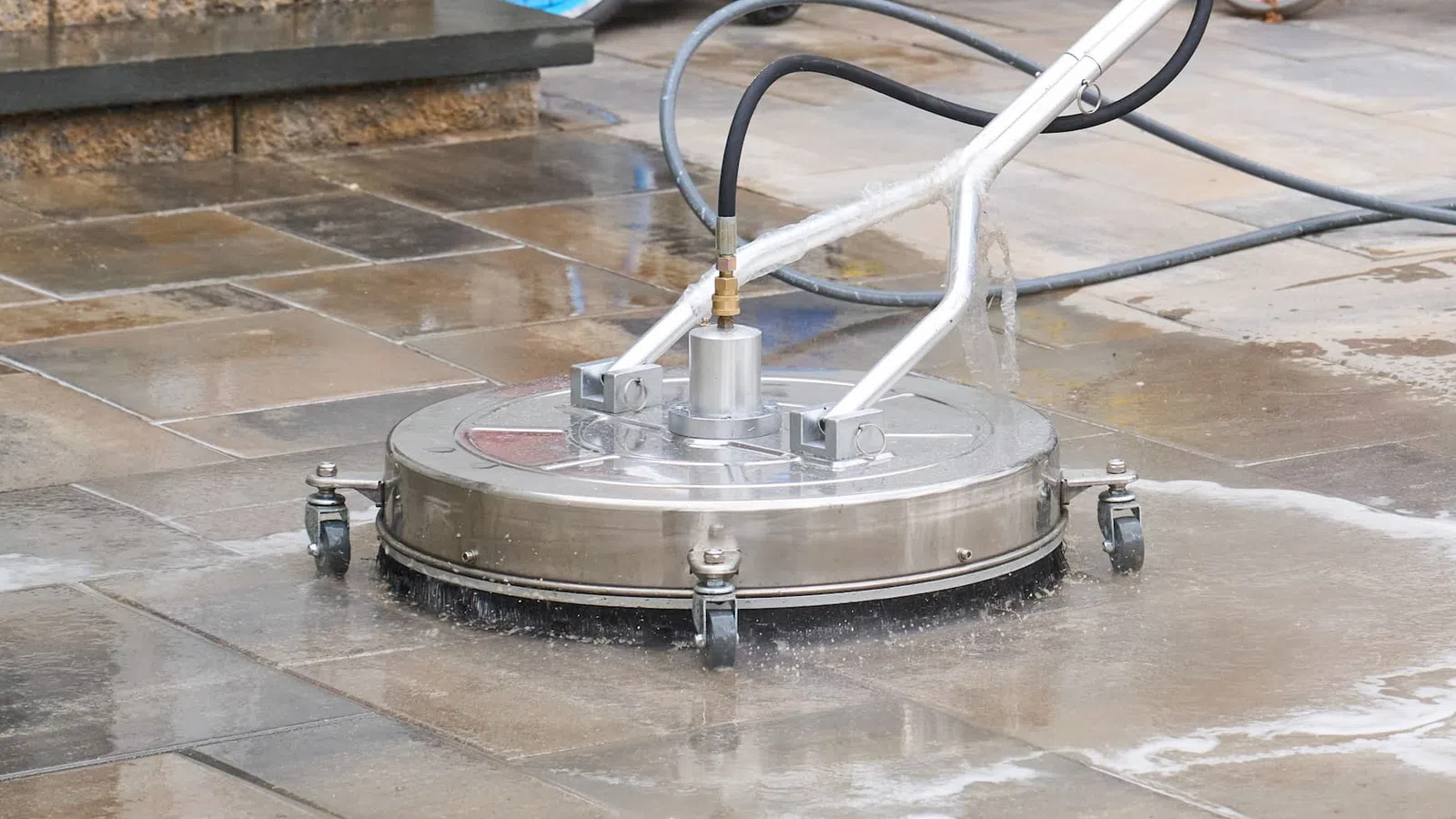 A stainless steel surface cleaner attached to a pressure washer cleaning wet outdoor tile flooring.