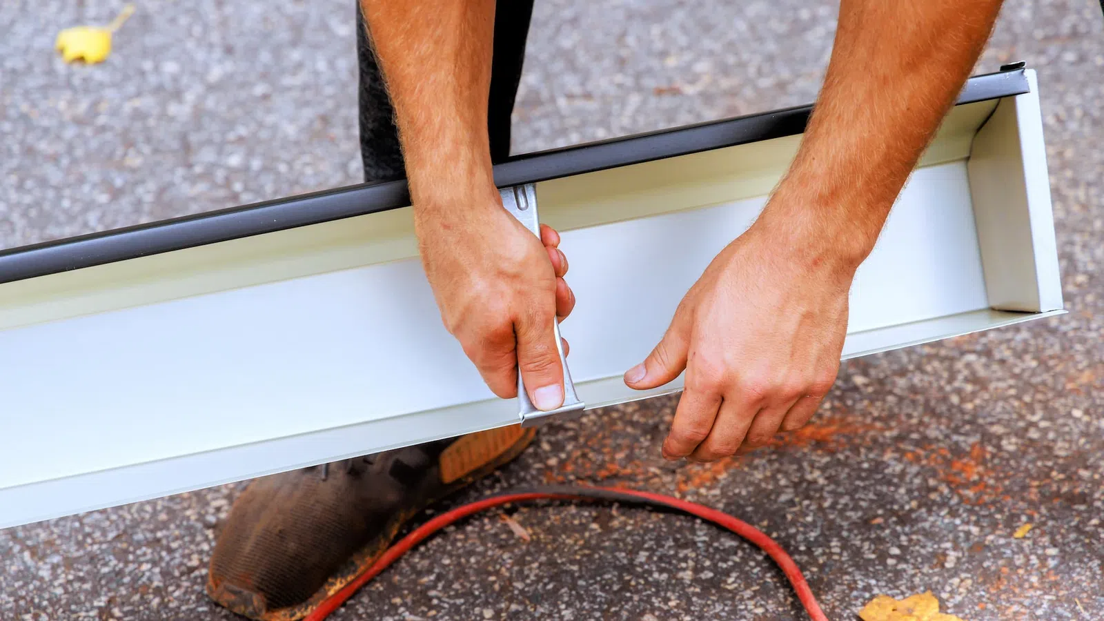 Person assembling a white metal light fixture outdoors, hands adjusting mounting bracket, brown shoe and red cord visible
