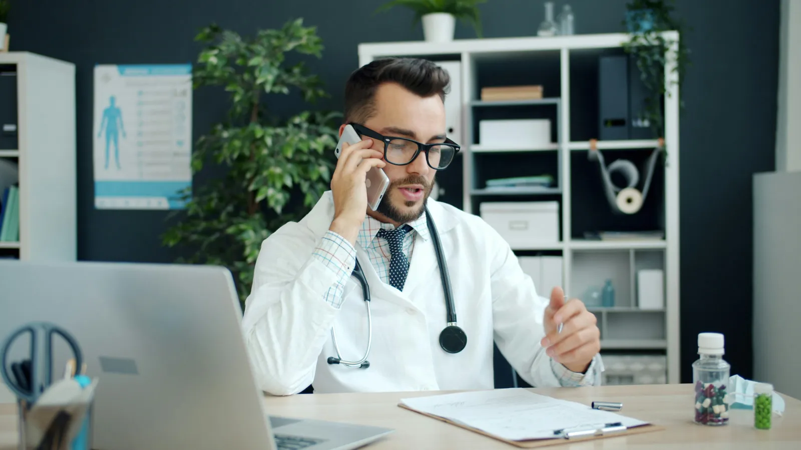 Male doctor in white coat talking on phone at desk with laptop and medical documents in office setting