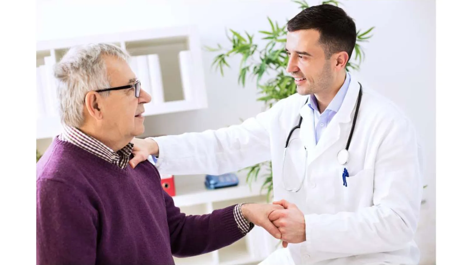 Doctor in white coat holding and reassuring elderly male patient in medical office with plant in background