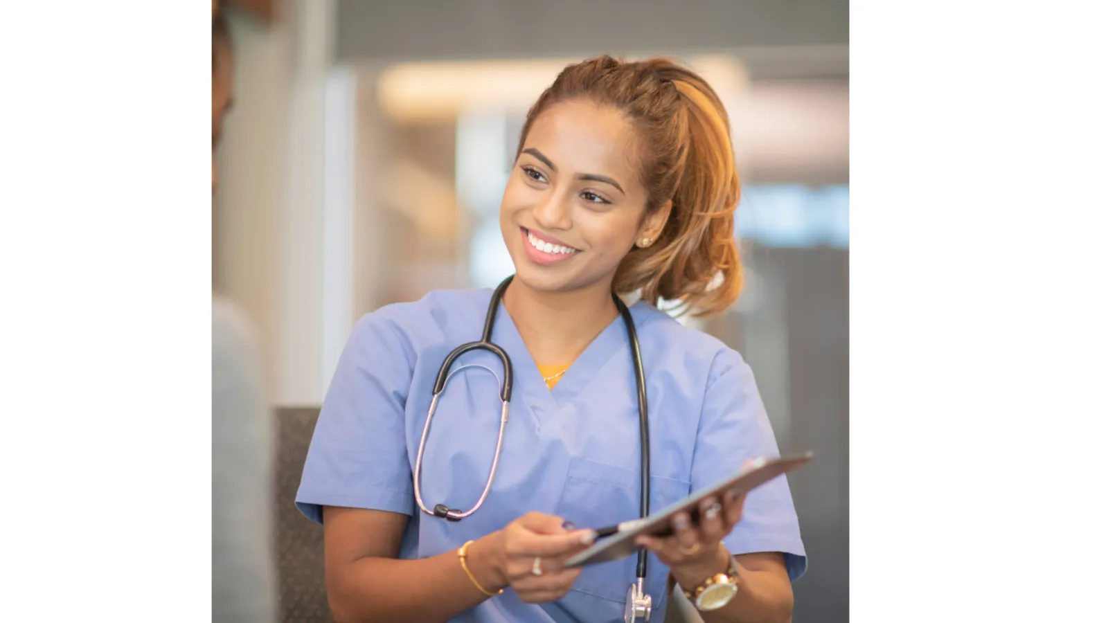 Smiling female healthcare professional in scrubs discussing with a patient, holding a tablet.