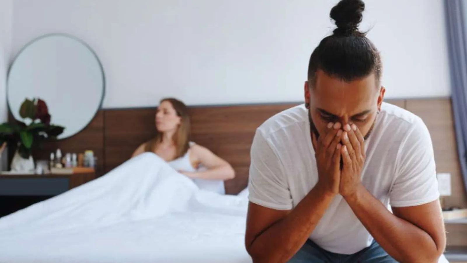 Man sitting on bed with head in hands, woman sitting on bed in background, expressing tension or sadness