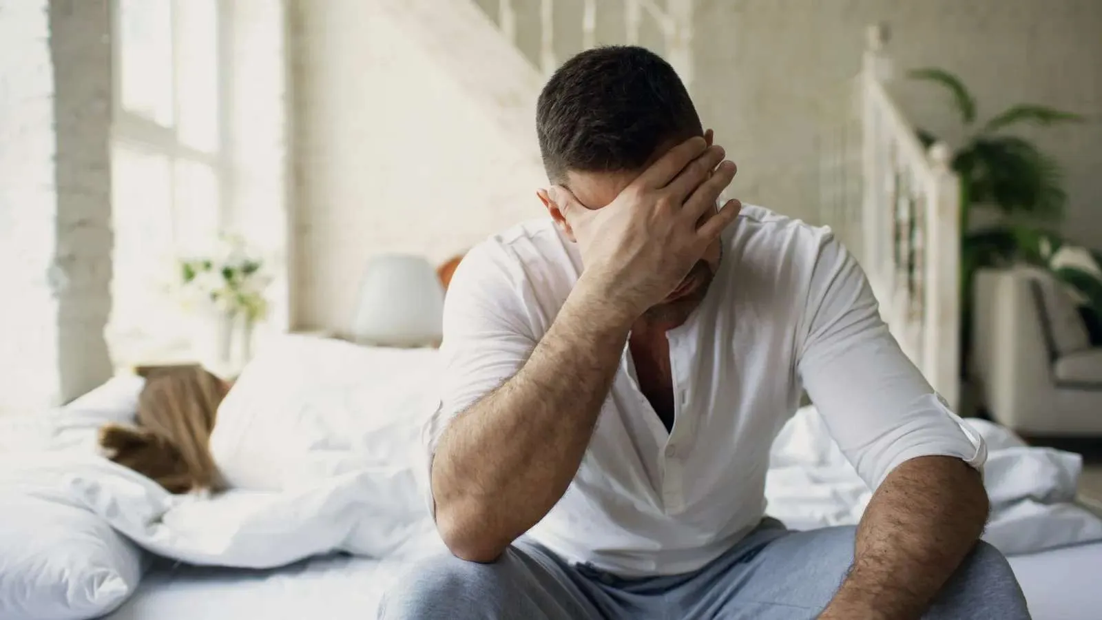 Man in white shirt sitting on bed with hand on forehead appearing stressed, woman sleeping in background.