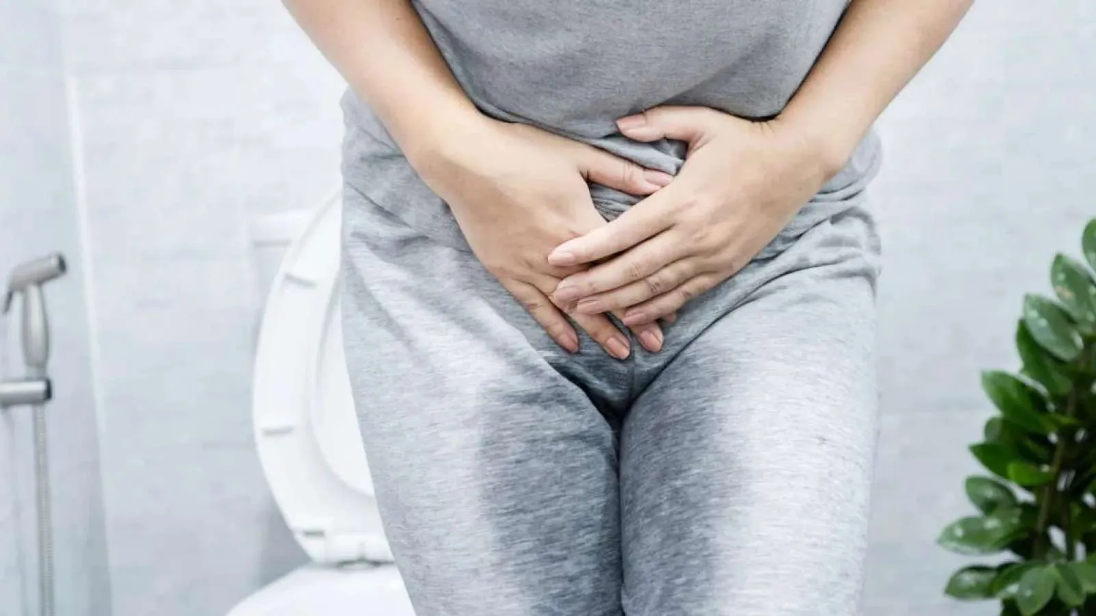 Person in gray clothes holding lower abdomen sitting on toilet, indicating urinary or bladder discomfort.