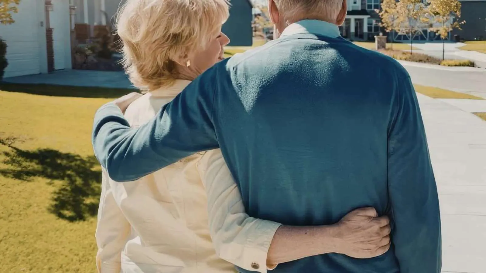 Elderly couple walking arm in arm outdoors in sunny neighborhood with houses and trees.