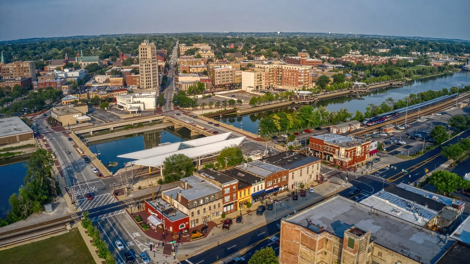 Aerial view of a riverfront city with bridges, historic buildings, train tracks, and green trees under a blue sky.