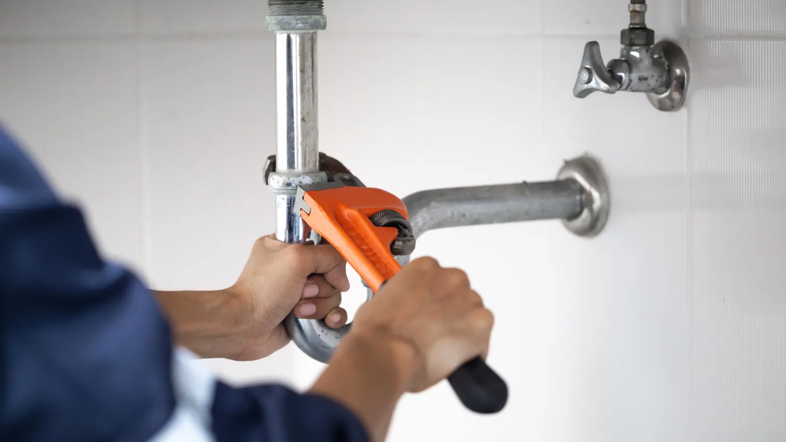 Plumber using an orange pipe wrench to tighten a metal pipe under a sink in a white tiled bathroom.
