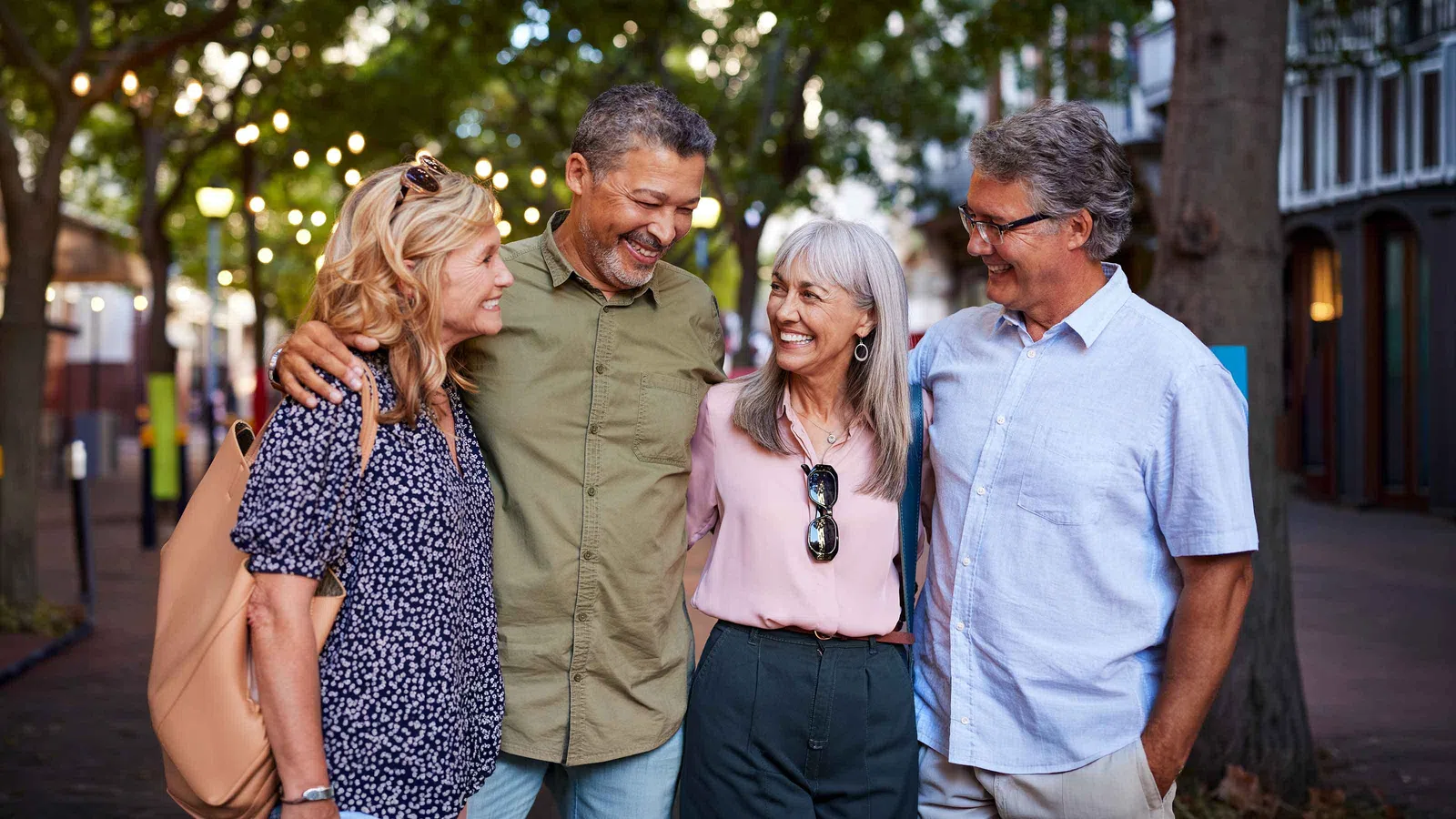 Happy mature friends smiling and walking together outdoors on a tree-lined street in casual attire.