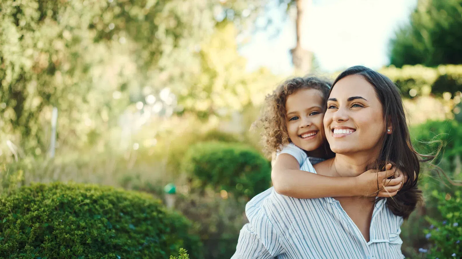 Smiling mother giving her daughter a piggyback ride outdoors in a sunny green park.