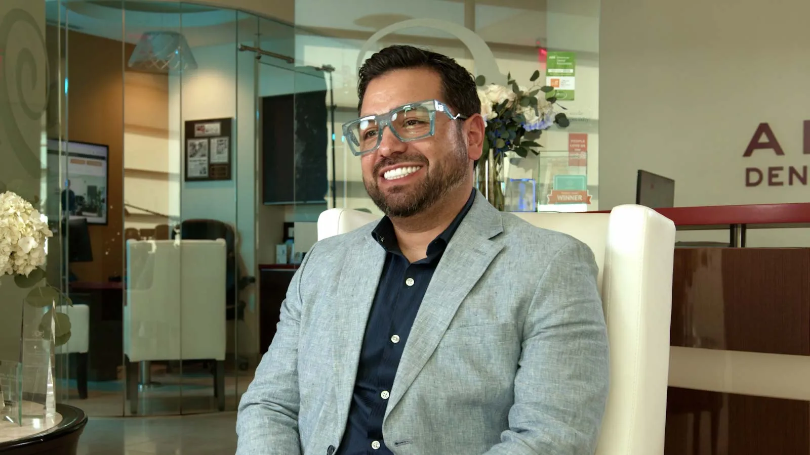 Smiling man wearing glasses and a gray blazer sitting in a modern office with glass walls and white flowers.