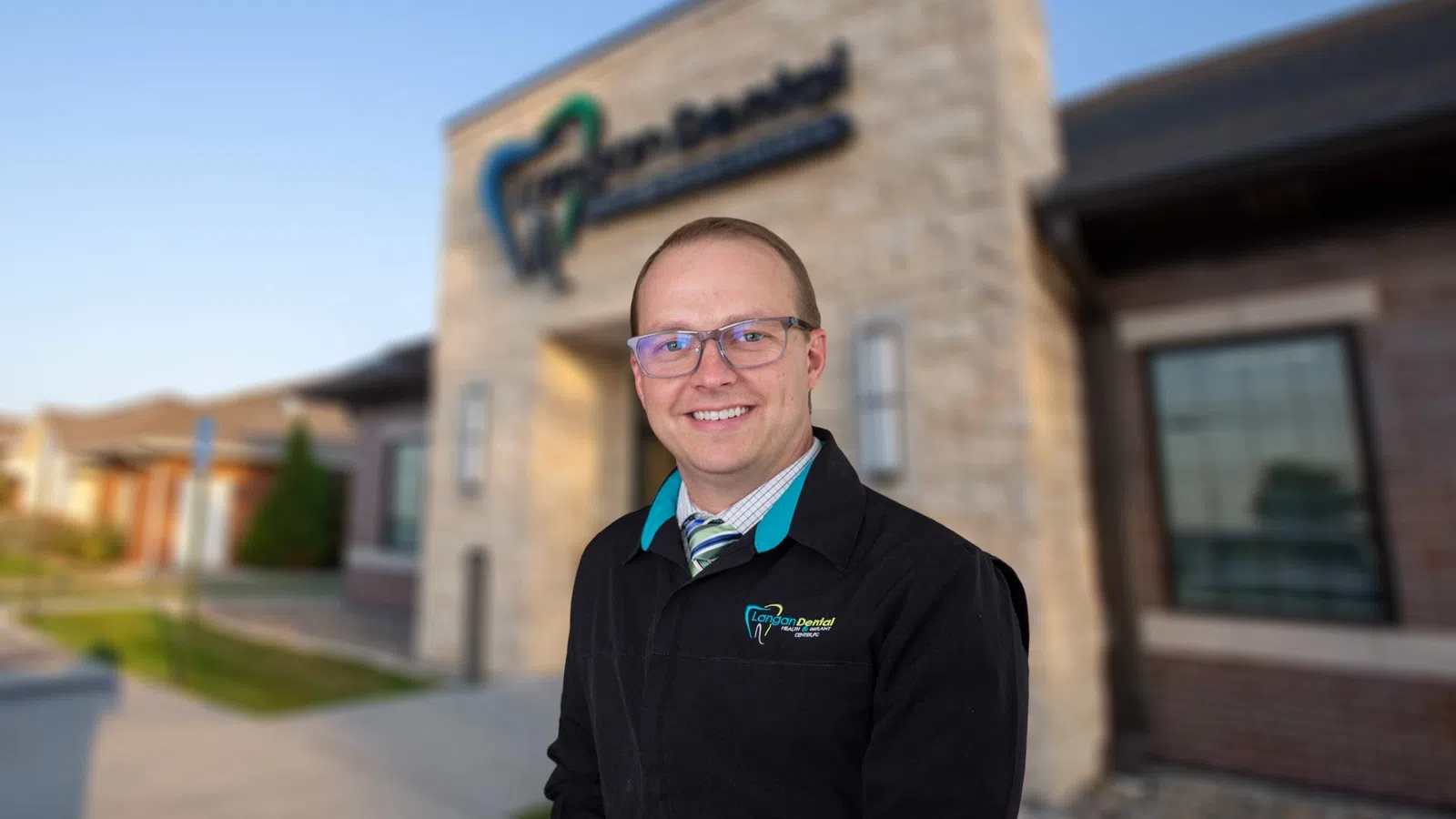 Smiling male dentist wearing glasses and a clinic jacket standing outside Canyon Dental clinic building.