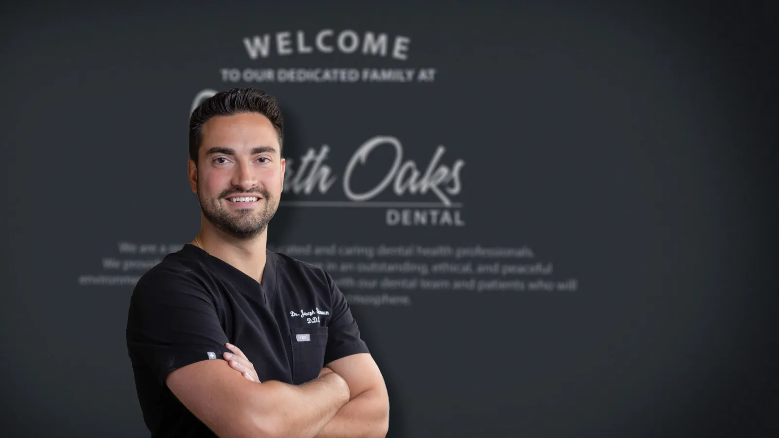 Smiling male dentist wearing black scrubs stands confidently with arms crossed in front of dental office sign.