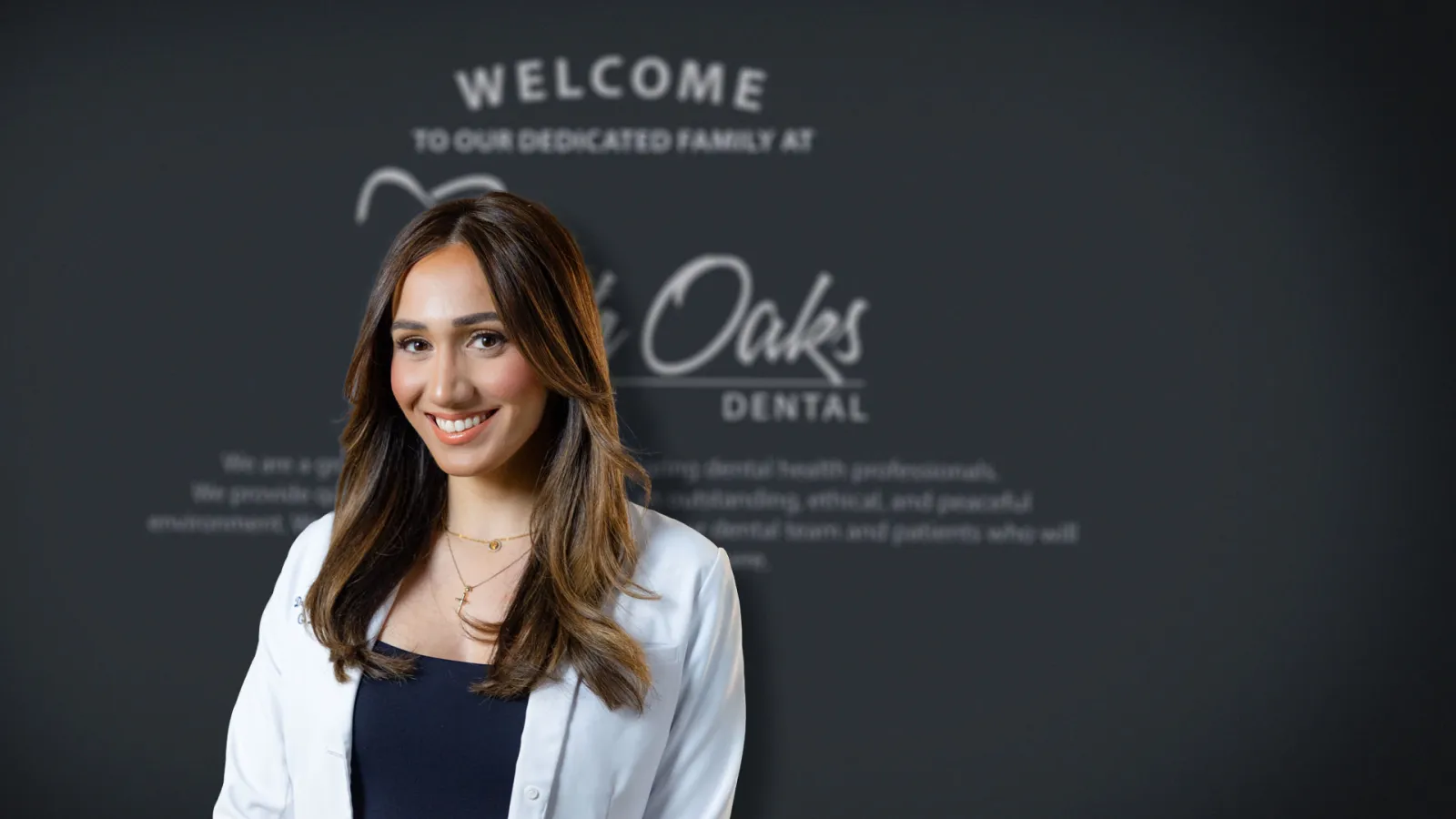 Smiling female dentist in white coat standing in front of a dental clinic welcome sign