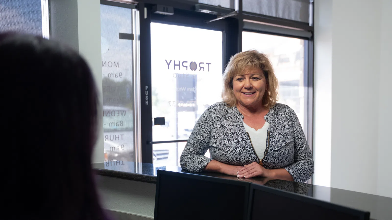 Smiling woman stands behind reception desk in bright office with glass door behind her showing business hours.