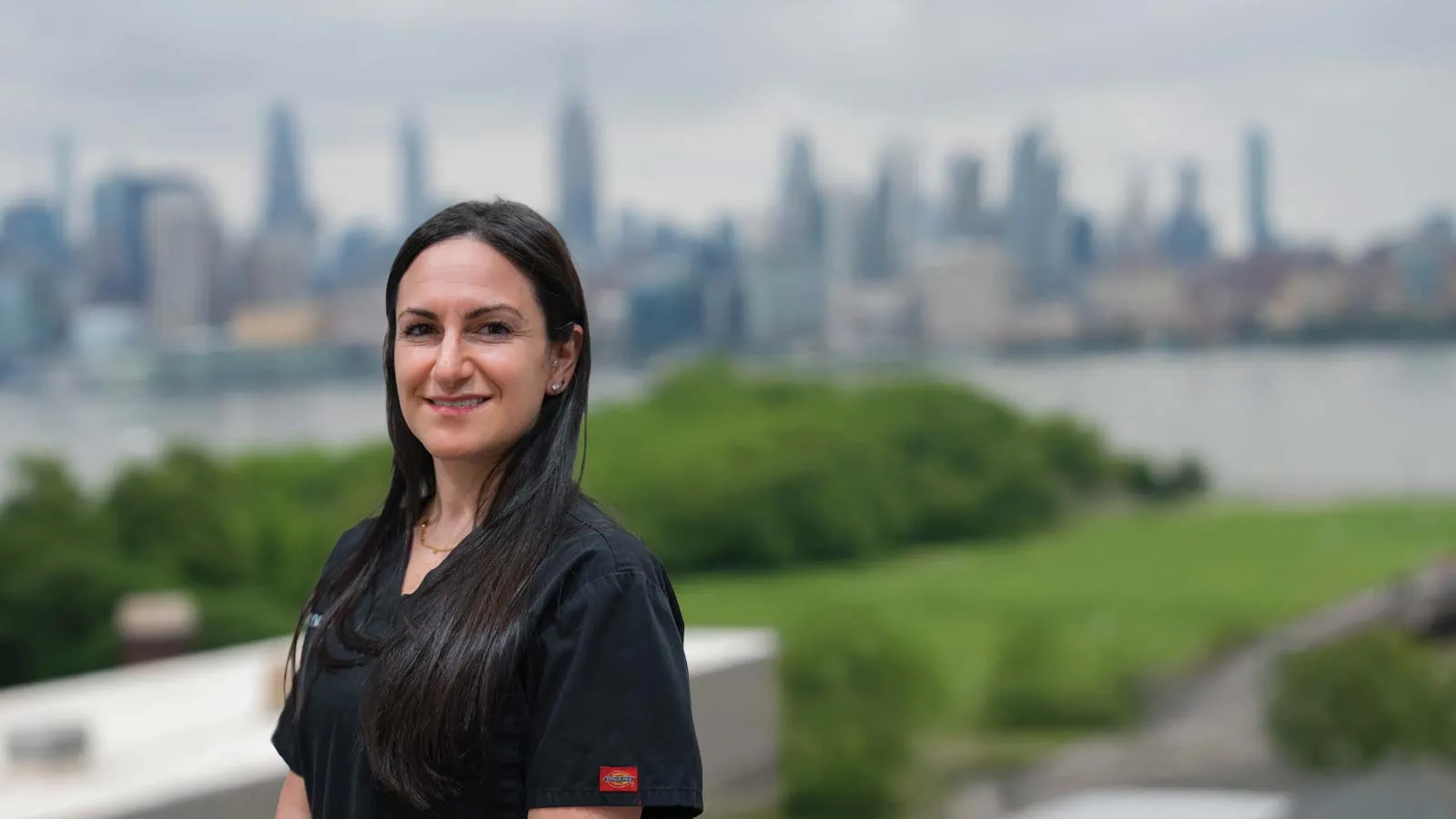 Smiling woman with long dark hair in black shirt standing outdoors with city skyline and green park in background.