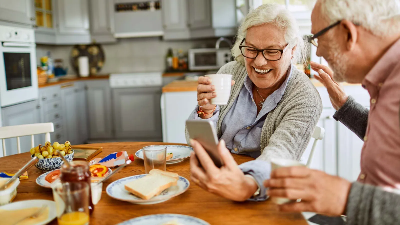 Senior couple enjoying breakfast together while looking at a smartphone and laughing in a cozy kitchen.