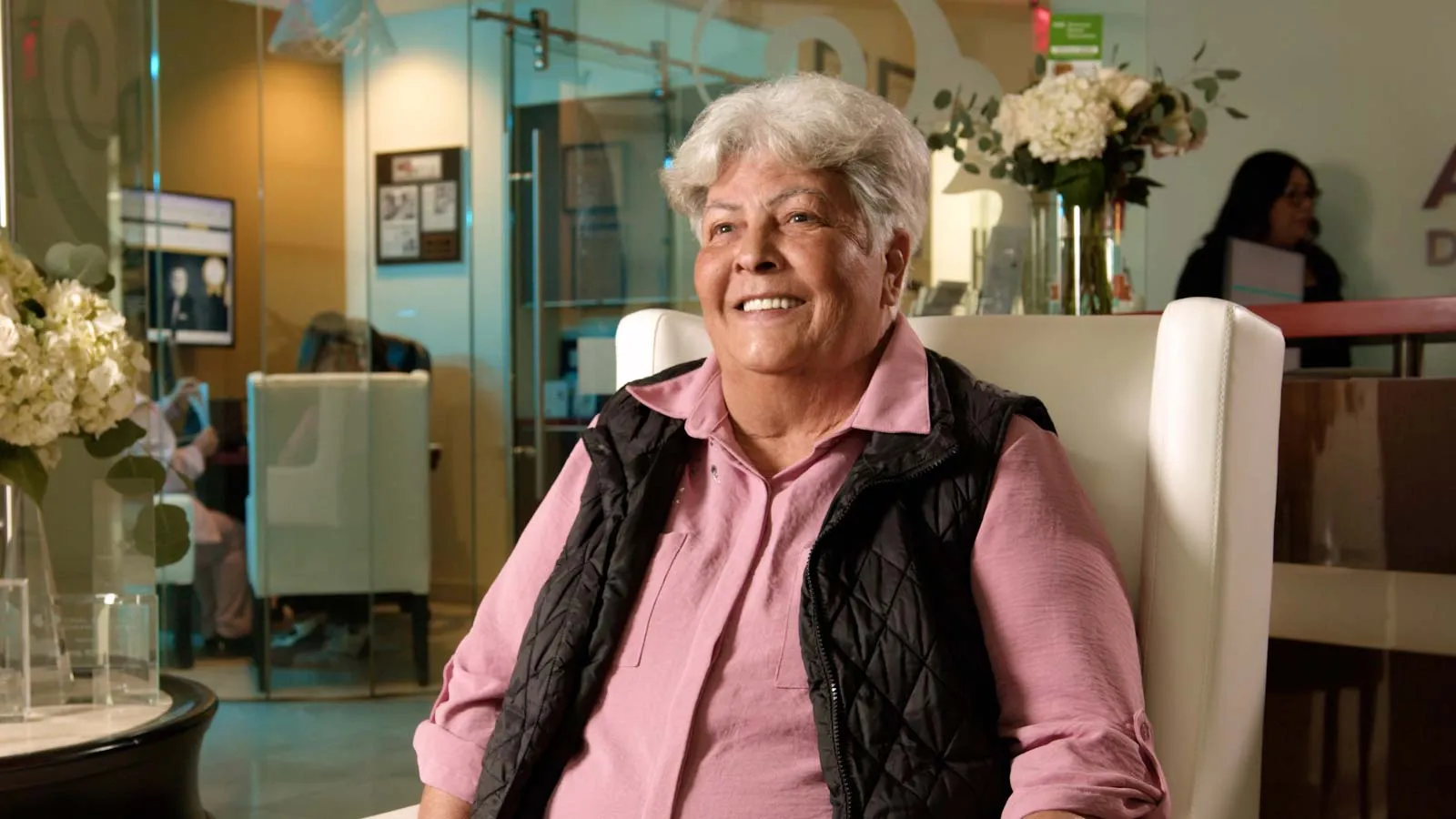 Smiling elderly woman with gray hair wearing a pink shirt and black vest seated in a bright modern room.