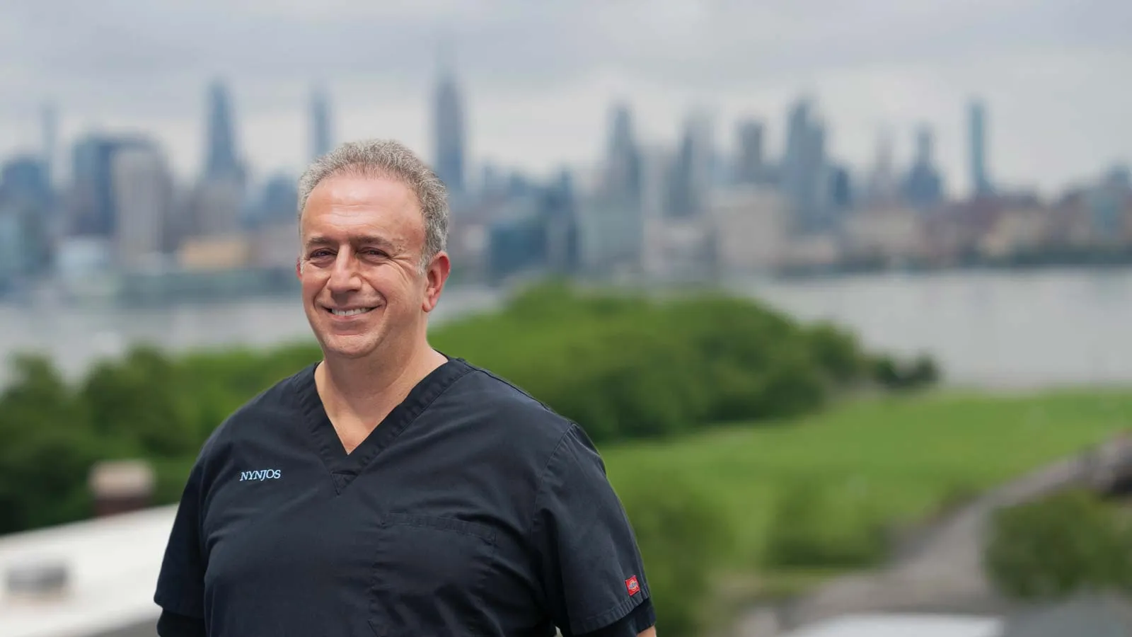 Smiling man in black medical scrubs standing outdoors with a city skyline and greenery in the background.